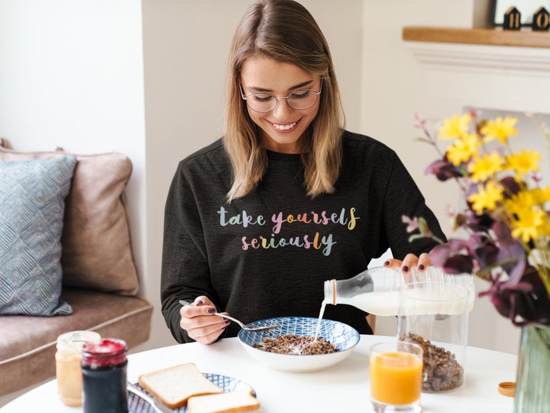 May include: A woman wearing a black sweatshirt with the text "take yourself seriously" in rainbow colors pours milk into a bowl of cereal.