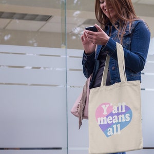 May include: A beige tote bag with a pink, blue, and purple heart that says "Y'all means all".