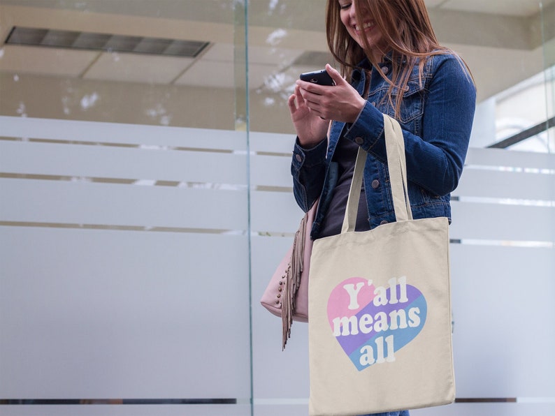 May include: A beige tote bag with a pink, blue, and purple heart that says "Y'all means all".