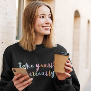 May include: A black sweatshirt with the text "take yourself seriously" in rainbow colors. The person is holding a brown paper cup and a smartphone.