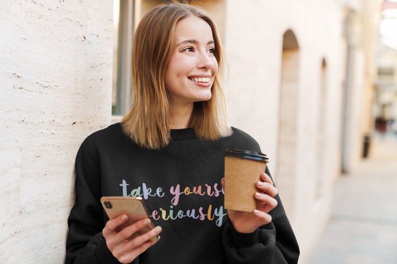 May include: A black sweatshirt with the text "take yourself seriously" in rainbow colors. The person is holding a brown paper cup and a smartphone.