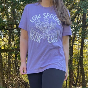 May include: A purple t-shirt with a white graphic of a book with flowers and the text "Low Spoons Book Club".