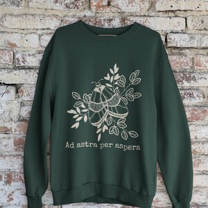Op de afbeelding: Een donkergroen sweatshirt met een witte afbeelding van twee motten op een tak met bladeren. De tekst "Ad astra per aspera" is onder de afbeelding gedrukt.