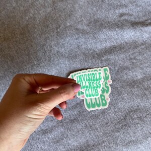 May include: A stack of green and white stickers with the text "Invisible Illness Club" in a retro font. The stickers have small pink hearts around the text.