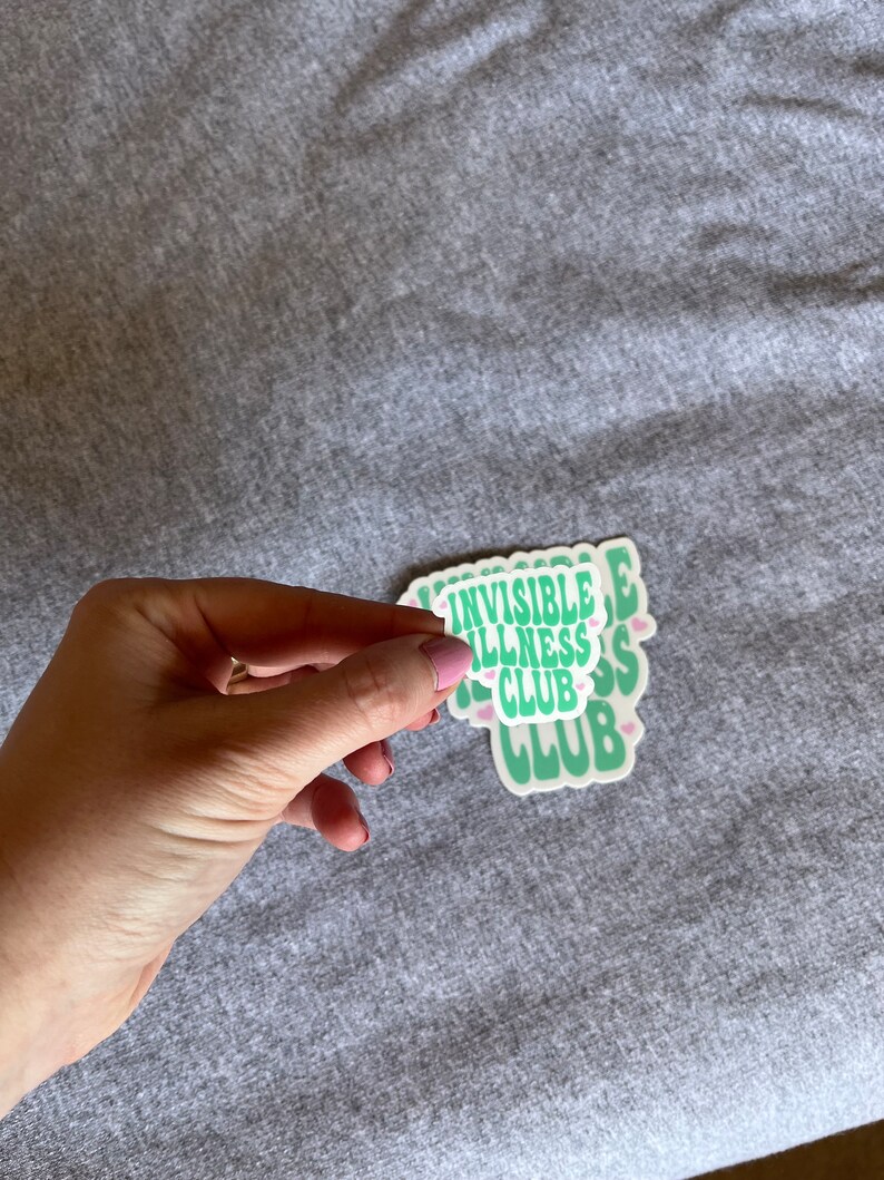 May include: A stack of green and white stickers with the text "Invisible Illness Club" in a retro font. The stickers have small pink hearts around the text.