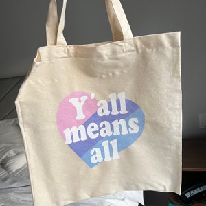 May include: A beige canvas tote bag with a pink, blue, and purple heart design. The heart has the text "Y'all means all" printed on it.