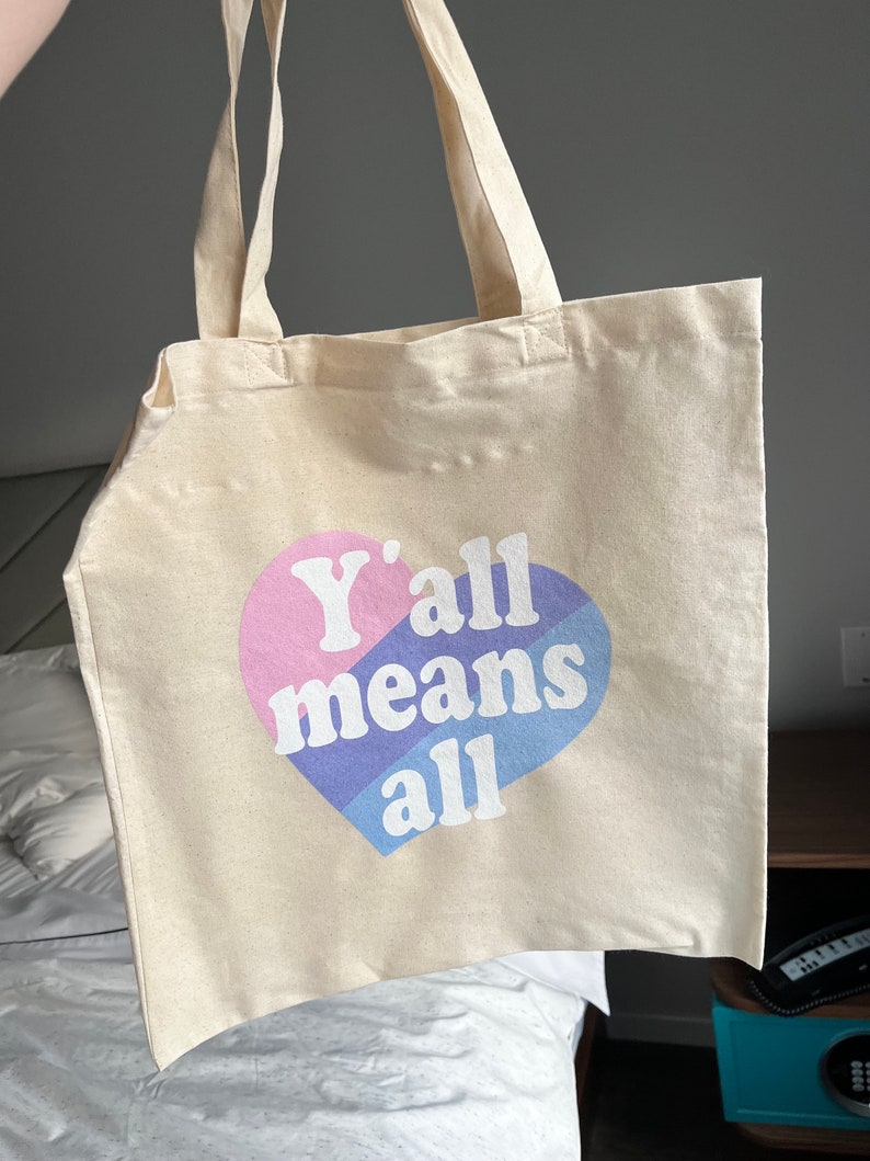 May include: A beige canvas tote bag with a pink, blue, and purple heart design. The heart has the text "Y'all means all" printed on it.