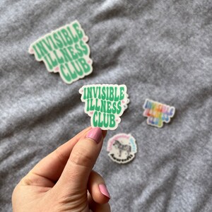 May include: A green sticker with the text "Invisible Illness Club" in a retro font. The sticker has two small pink hearts on either side of the text.