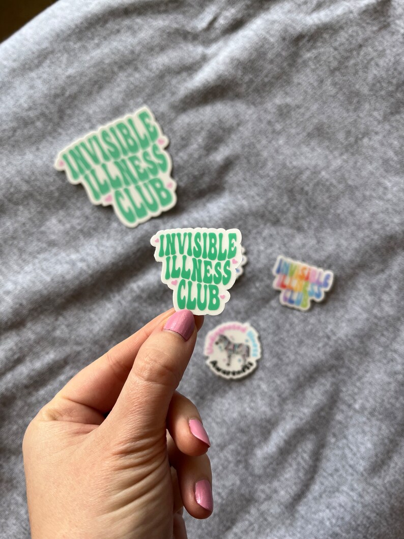 May include: A green sticker with the text "Invisible Illness Club" in a retro font. The sticker has two small pink hearts on either side of the text.