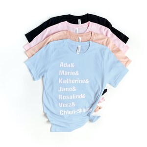May include: A light blue t-shirt with white text that reads "Ada & Marie & Katherine & Jane & Rosalind & Vera & Chien-Shiung". The shirt is folded and stacked on top of other t-shirts in pink, peach, and black.