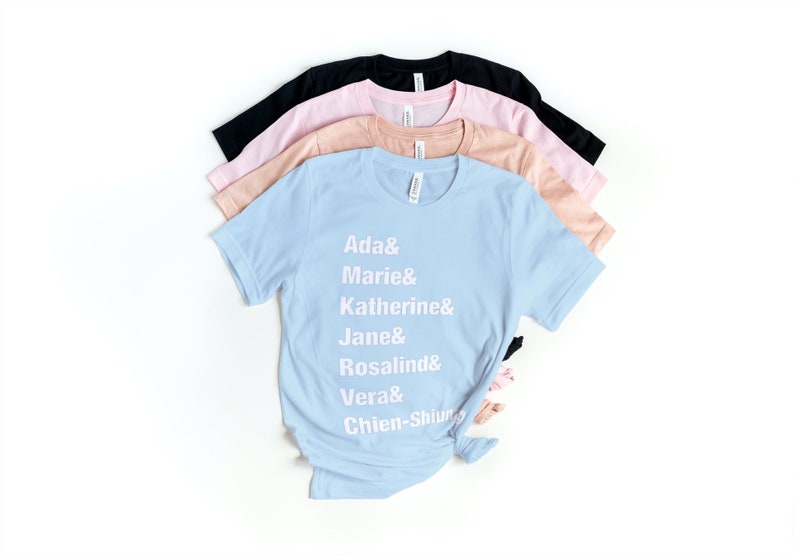 May include: A light blue t-shirt with white text that reads "Ada & Marie & Katherine & Jane & Rosalind & Vera & Chien-Shiung". The shirt is folded and stacked on top of other t-shirts in pink, peach, and black.