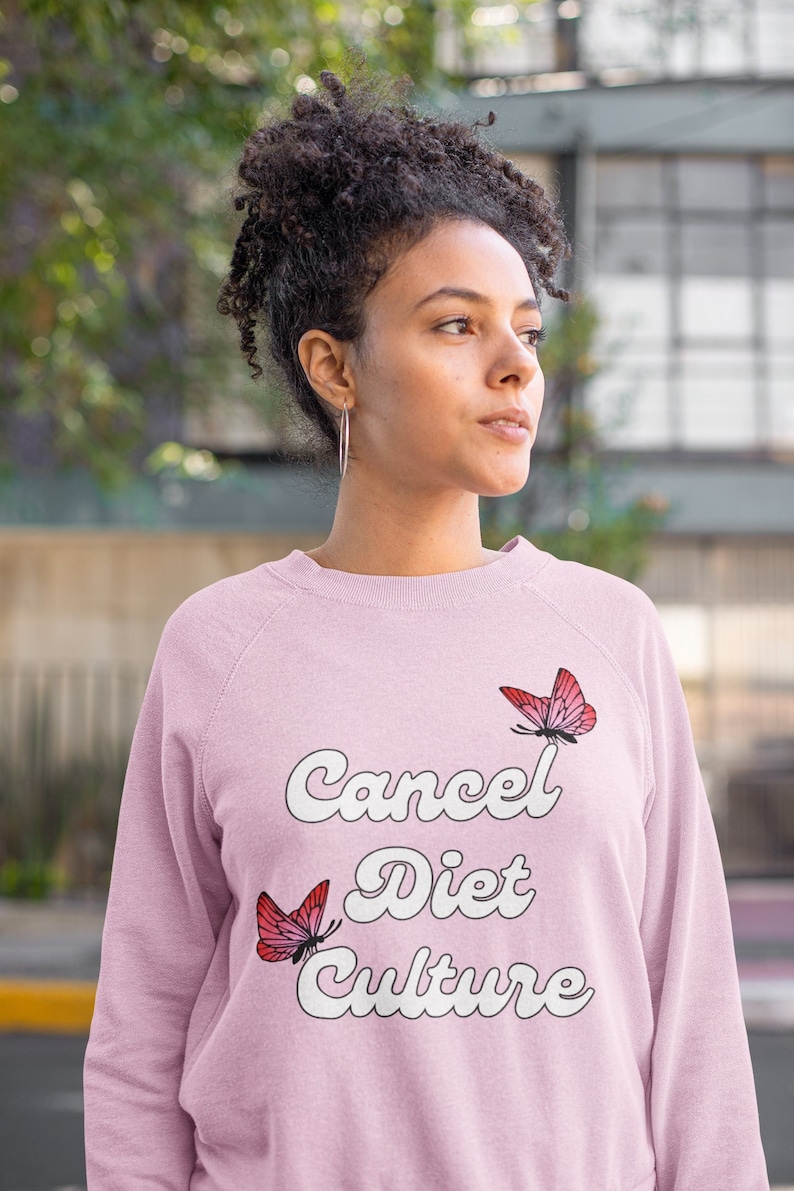 Pu&ograve; includere: Una felpa rosa con il testo "Cancel Diet Culture" in bianco con un font retr&ograve;. Due farfalle rosa sono sulla felpa, una sopra il testo e una sotto.