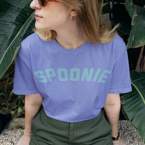 A woman is wearing a purple oversized tee shirt in the color black that says in teal letters "SPOONIE".