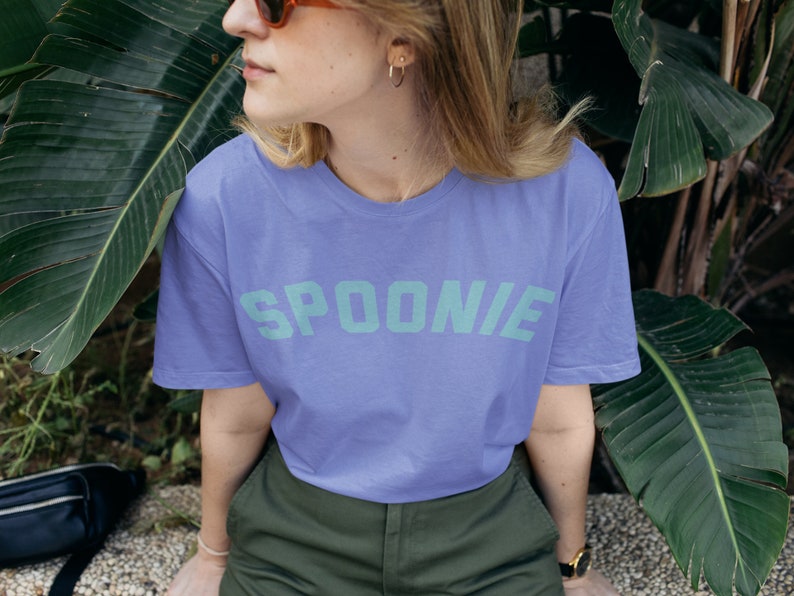 A woman is wearing a purple oversized tee shirt in the color black that says in teal letters "SPOONIE".