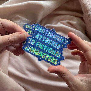 May include: A blue holographic sticker with the text "Emotionally Attached to Fictional Characters" with a heart symbol on either side of the text.