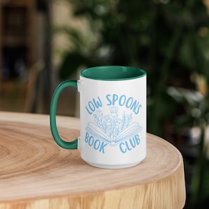 May include: White ceramic mug with a teal rim and handle. The mug has a blue design of an open book with flowers growing out of it. The text "Low Spoons Book Club" is printed on the mug.
