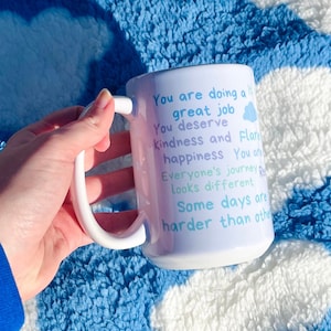 May include: White ceramic mug with a light blue and pink ombre design. The mug has a quote that reads: "You are doing a great job. You deserve kindness and happiness. Everyone's journey looks different. Some days are harder than others."