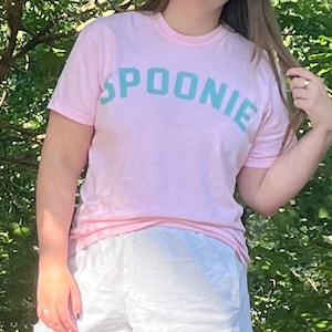 May include: A pink t-shirt with the word "SPOONIE" printed in teal blue letters.