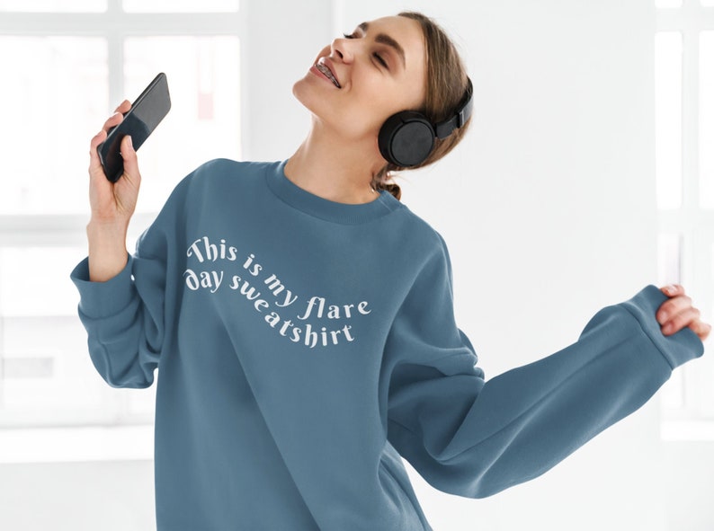 A young woman with a blue teal crewneck sweatshirt on that has wavy text that says "This is my flare day sweatshirt".