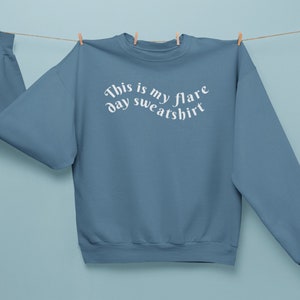 A blue teal colored crewneck sweatshirt on that has wavy text that says "This is my flare day sweatshirt" is being held up by clothes pins on a clothes line