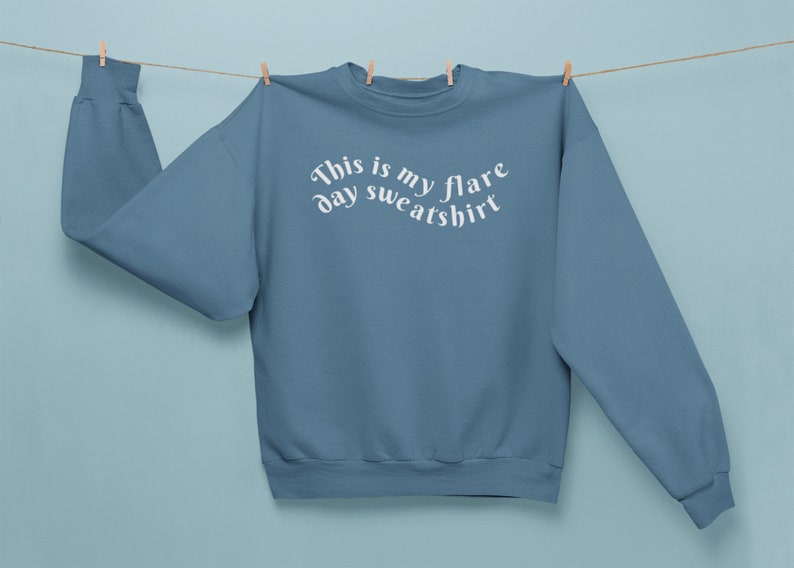 A blue teal colored crewneck sweatshirt on that has wavy text that says "This is my flare day sweatshirt" is being held up by clothes pins on a clothes line