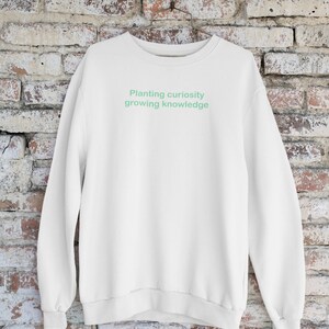 Puede incluir: Sudadera blanca con un gr&aacute;fico de texto verde que dice "Planting curiosity growing knowledge".