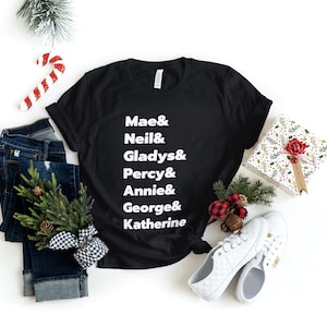 May include: Black t-shirt with white text that reads "Mae & Neil & Gladys & Percy & Annie & George & Katherine". The shirt is laid flat on a white surface with a pair of jeans, a small potted tree, a candy cane, a gift box, and a pair of white sneakers.