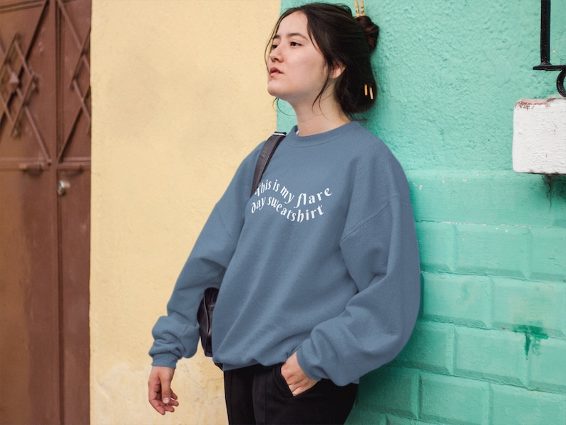 A young woman leaning against a light green wall with a steel blue teal crewneck sweatshirt on that has wavy text that says "This is my flare day sweatshirt". She is looking away and looks kind of tired.