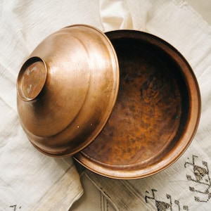 May include: A copper bowl with a lid, partially open, on a white cloth with a patterned design. The bowl and lid have a warm, metallic sheen, suggesting a handcrafted item.