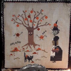 May include: A cross-stitch embroidery featuring a witch holding a basket of apples, a tree with a face, and a black cat. The text "SPLASH" and "BOOM" are also included in the design.