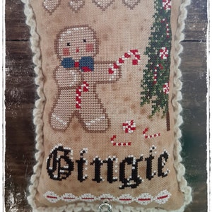 Può includere: Un cuscino beige con un bordo all'uncinetto presenta un omino di pan di zenzero che tiene un bastoncino di zucchero e un albero di Natale verde con bastoncini di zucchero. Il cuscino è ricamato con la parola "Jingle".