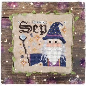 Può includere: Un ricamo a punto croce quadrato su tessuto beige raffigurante un mago con una lunga barba bianca, un cappello blu con stelle gialle e un bastone con una sfera azzurra. Il testo "MERLIN" e "Sep" è ricamato in nero. Bordo rifinito con spago.