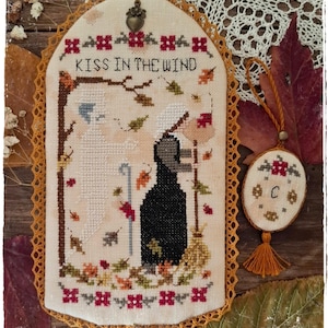 Può includere: Opera d'arte a punto croce con la scritta "KISS IN THE WIND" e un bordo decorativo. L'opera presenta una strega e un fantasma con foglie autunnali. Viene mostrato anche un ornamento più piccolo abbinato con un nappa.