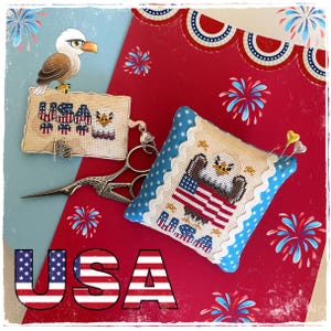 May include: Cross-stitch USA-themed pincushion and needle holder with an American flag and eagle design. The pincushion has a blue star border. A pair of scissors and a small American flag charm are also present. The word "USA" is displayed in red, white, and blue.