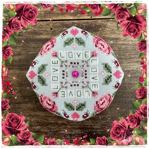 Puede incluir: Un diseño de punto de cruz cuadrado azul claro con la palabra "LOVE" bordada en blanco. El diseño está rodeado de rosas rosas y hojas verdes. Una cuenta rosa está en el centro del diseño.