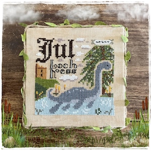 Può includere: Un ricamo a punto croce quadrato su tessuto beige raffigurante una creatura blu nell'acqua, un albero verde, un edificio marrone e il testo "Jul Loch Ness" e "Nessy". Il pezzo è incorniciato da un bordo in corda rustica con foglie in tessuto.