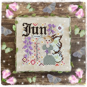 Può includere: Un disegno a punto croce su tessuto beige presenta la parola "Jun" in lettere marroni scure, con un'illustrazione di una fata. Il disegno include fiori viola, foglie verdi e la parola "Fairy". Farfalle e fiori rosa circondano il disegno.
