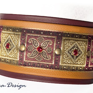 May include: A brown leather dog collar with a gold and burgundy patterned fabric insert. The collar is decorated with gold studs and red rhinestones. "Luca-Design" is written in blue script on the bottom of the image.