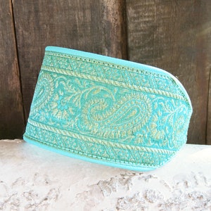May include: A turquoise fabric cuff with an ornate paisley design in gold and light green. The cuff is resting on a white surface with a wooden background.