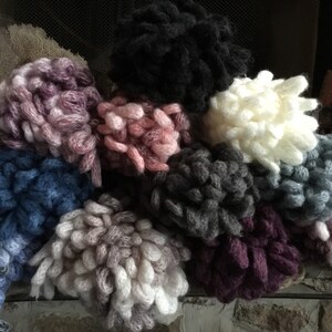 Puede incluir: Una colecci&oacute;n de diez ovillos de lana esponjosos en varios colores, incluyendo rosa, morado, gris, azul y blanco. Los ovillos de lana est&aacute;n dispuestos en una pila sobre una superficie texturizada.