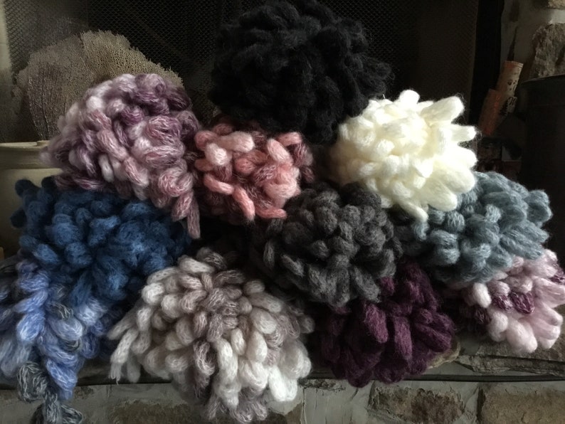 Puede incluir: Una colecci&oacute;n de diez ovillos de lana esponjosos en varios colores, incluyendo rosa, morado, gris, azul y blanco. Los ovillos de lana est&aacute;n dispuestos en una pila sobre una superficie texturizada.