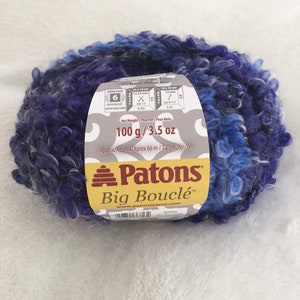 Op de afbeelding: Een bol Patons Big Bouclé garen in een blauwe en paarse kleurstelling. Het garen is super dik en heeft een pluche textuur. Het etiket geeft aan dat het garen 20% mohair bevat en 100 gram of 3,5 ounce weegt. Het etiket bevat ook de naaldgrootte aanbevelingen voor het breien met dit garen.