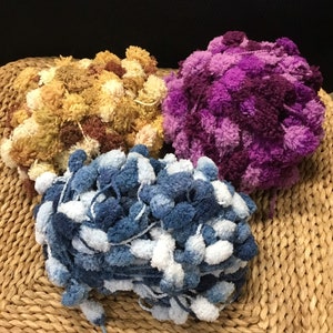 May include: Three skeins of yarn with pom poms. One skein is a mix of brown and beige, one is purple, and one is blue and white.