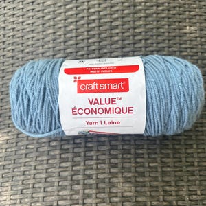 May include: A ball of Craft Smart Value yarn in a pale blue shade. The label displays "VALUE ÉCONOMIQUE Yarn | Laine" and the Craft Smart logo. The yarn is 100% acrylic, ideal for knitting and crochet. The label also says "PATTERN INCLUDED MOTIF INCLUS".