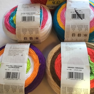 May include: Five balls of yarn in various colours, including pink, white, blue, orange, purple, brown, and green. Each ball has a label with the name of the yarn colour and a barcode. The yarn is for crocheting or knitting.