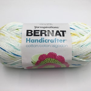 May include: A ball of Bernat Handicrafter cotton yarn in a white, yellow, blue, and green colourway. The yarn label reads "Bernat Handicrafter cotton/coton/algodón" and features a crocheted flower.