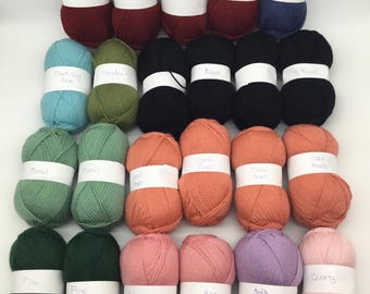 May include: Twenty-four skeins of yarn in various colors, including bright red, duck egg blue, indigo, meadow green, black, coral peach, basil green, pine green, rose, soft orchid, and quartz. Each skein has a white label with the color name printed on it.