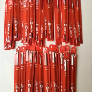May include: A collection of 20 red and silver knitting needles in various sizes, ranging from 1.00 mm to 5.0 mm. The needles are packaged individually in clear plastic sleeves with red labels that say "AERO".