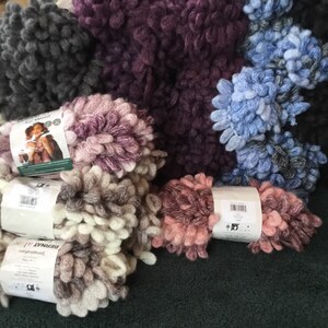 Puede incluir: Surtido de hilo Bernat Blanket en varios colores, incluyendo blanco, gris, morado, rosa y azul. El hilo es de estilo esponjoso y texturizado.