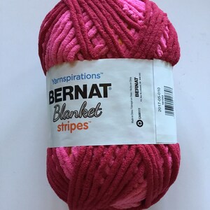 May include: A ball of pink and red yarn with a white label that reads "Yarnspirations&trade; BERNAT&reg; Blanket stripes&trade;". The label also includes the text "Made in China/Fabriqu&eacute; en Chine/Hecho en China", "CA00111", "BERNAT", "PO Box 40, Listowel, ON N4W 3J0", and "2017-05-010".